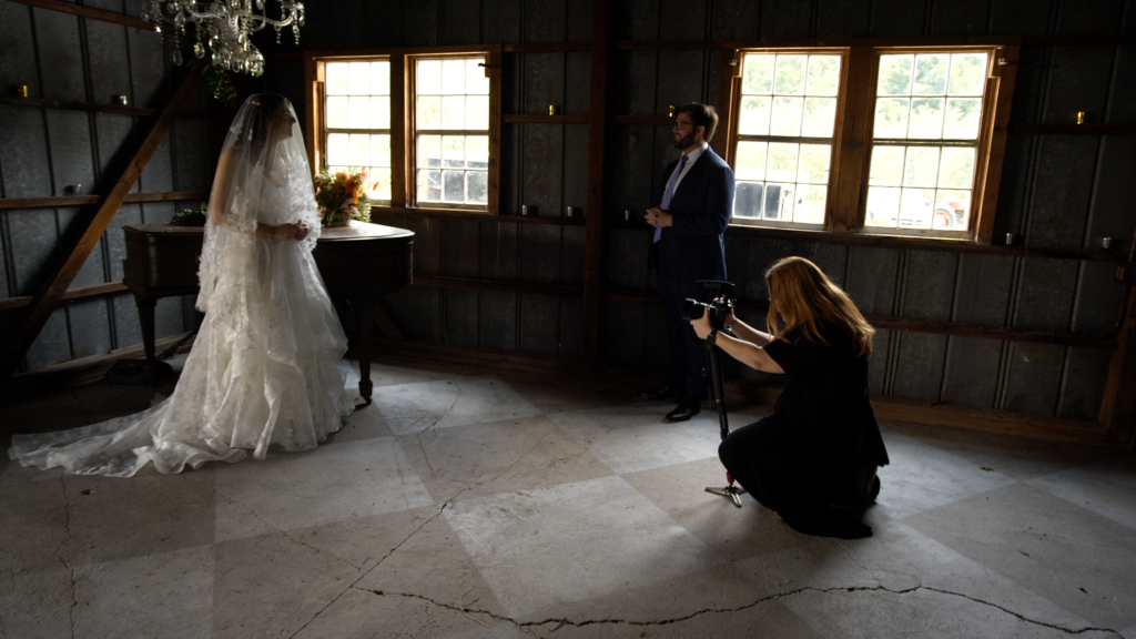 A bride in white poses in the window light of a rustic space while videographer, dressed in black, crouches down with a camera.